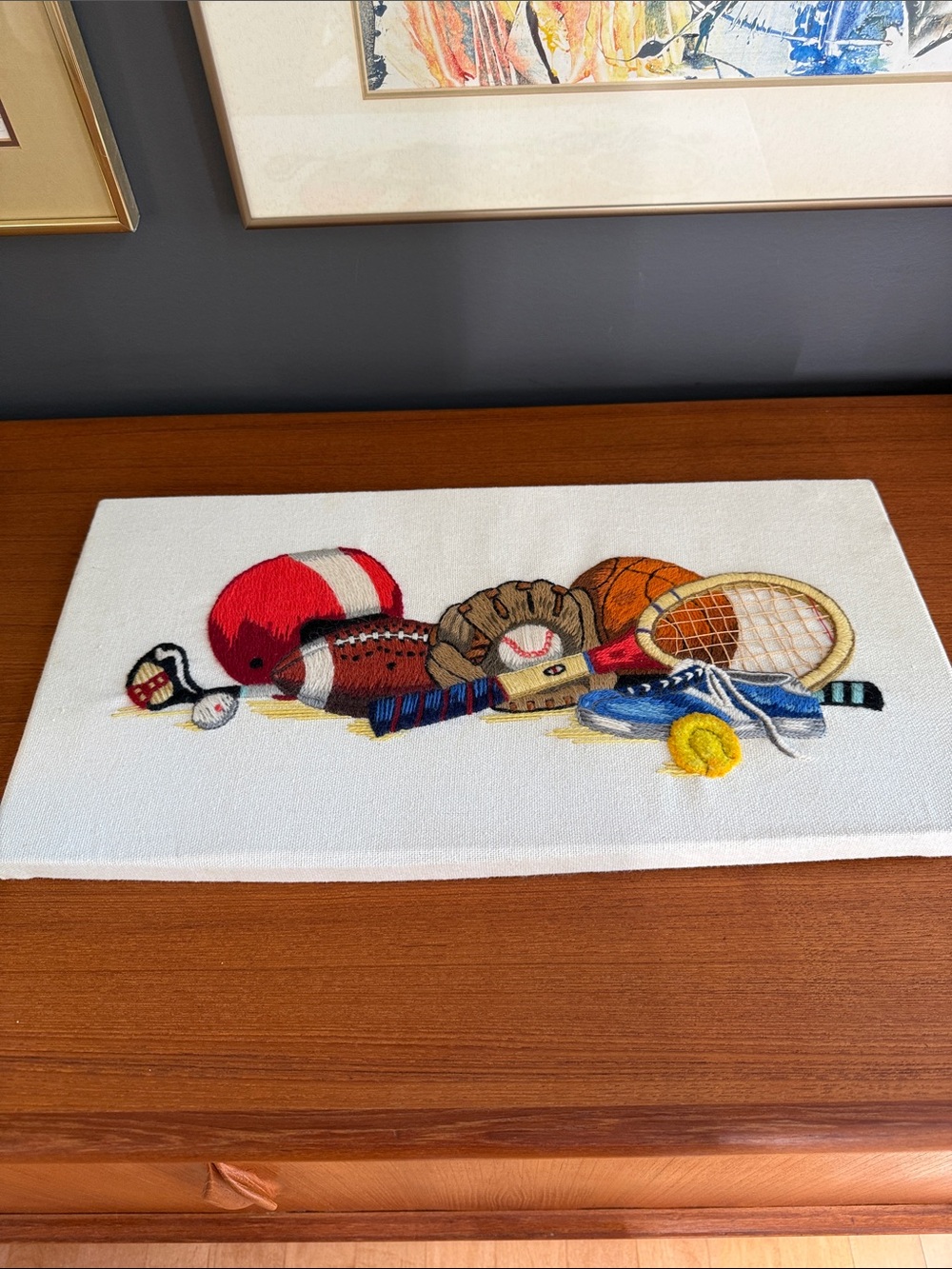 1976 crewel embroidery wall hanging features a stacked sports equipment